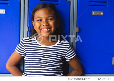 In school, young biracial female student leaning against blue lockers, smiling outdoors 116308978