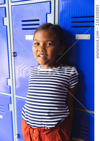 In school, young biracial girl standing by blue lockers, wearing striped shirt 116308983