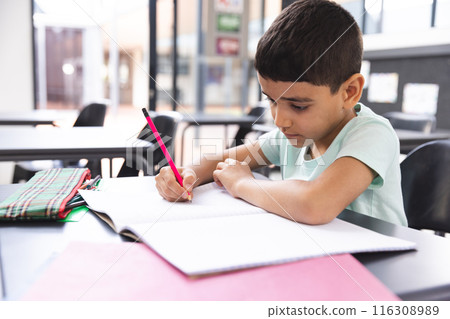 In school, young biracial boy focusing on writing in a notebook in the classroom 116308989