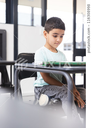 In school, young biracial male student sitting at a desk in a classroom, looking at a tablet 116308998