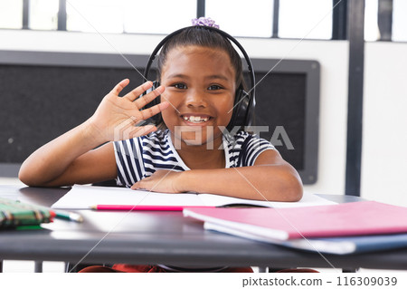 In school, young biracial girl wearing headphones waving at camera in classroom 116309039