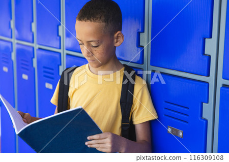Biracial boy reads a book by his school locker 116309108