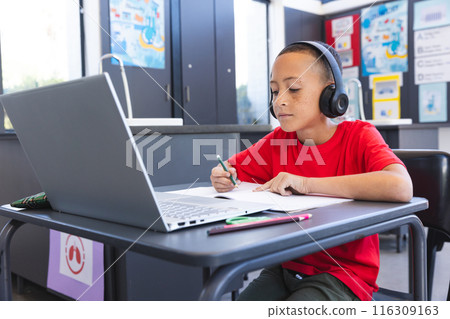 Biracial boy studies intently in a school classroom using a laptop, with copy space 116309163