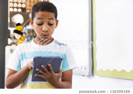 Biracial boy looks surprised at a tablet in a bright room at school 116309189