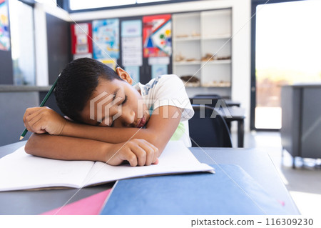 Exhausted biracial boy sleeps on desk in bright classroom. Exhausted biracial boy sleeps on desk in bright classroom. 116309230