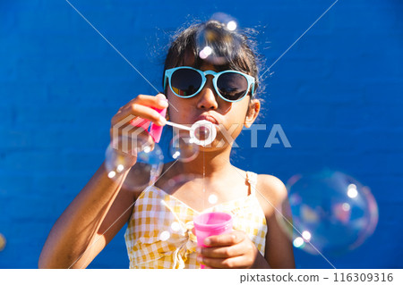 Biracial girl wearing sunglasses and a sundress enjoys blowing bubbles outdoors Biracial girl wearing sunglasses and a sundress enjoys blowing bubbles outdoors 116309316