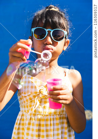 Biracial girl wearing sunglasses and a sundress blows bubbles outdoors 116309317