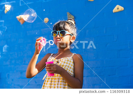 Biracial girl wearing sunglasses and a sundress enjoys blowing bubbles outdoors, with copy space 116309318