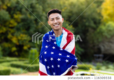 Portrait of happy asian man covered with flag of usa in garden Portrait of happy asian man covered with flag of usa in garden 116309338