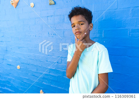 Biracial boy stands thoughtfully against a blue wall with copy space 116309361