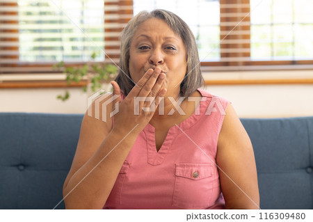 Biracial senior woman on blue couch blowing kiss during video call from home Biracial senior woman on blue couch blowing kiss during video call from home 116309480