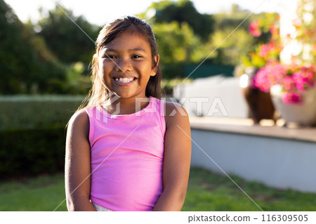 Outdoors, young biracial girl smiling brightly while standing in sunlit garden Outdoors, young biracial girl smiling brightly while standing in sunlit garden 116309505