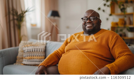 Fat lazy african american man on sofa at home wearing orange warm sweater. Disappointment and gain. 116309512