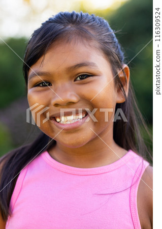 Outdoors, young biracial girl smiling brightly, wearing a pink top with greenery 116309524