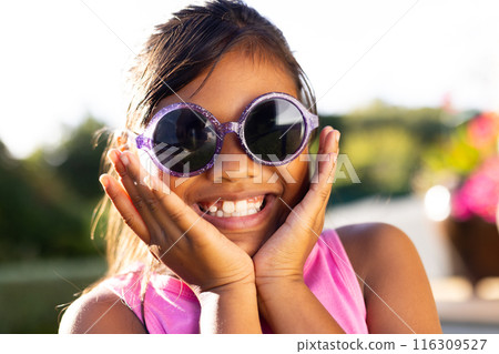 Outdoors, young biracial girl smiling brightly while wearing large, round sunglasses 116309527