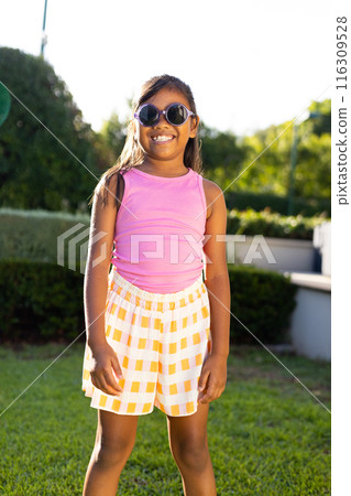 Young biracial girl in sunglasses, pink top, smiling brightly on grass outdoors 116309528