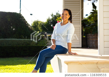 Outdoors, biracial woman in her late thirties sitting on porch steps, smiling warmly Outdoors, biracial woman in her late thirties sitting on porch steps, smiling warmly 116309533
