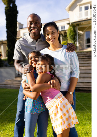 Outdoors, diverse family of four is smiling and hugging, in front of home 116309548
