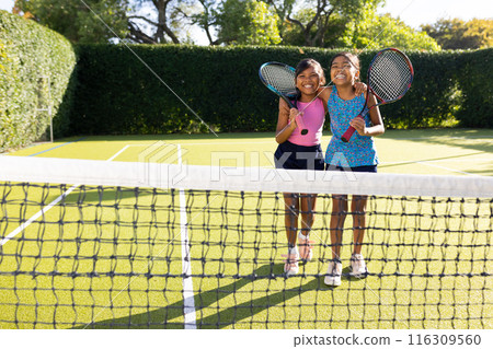Outdoors, two biracial young sisters smiling, holding tennis rackets on tennis court 116309560