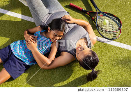 Outdoors, biracial mother and child lie on grass, smiling and embracing after tennis 116309597