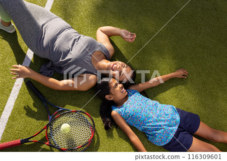 Outdoors biracial mother and daughter lying on grass with tennis rackets, smiling 116309601