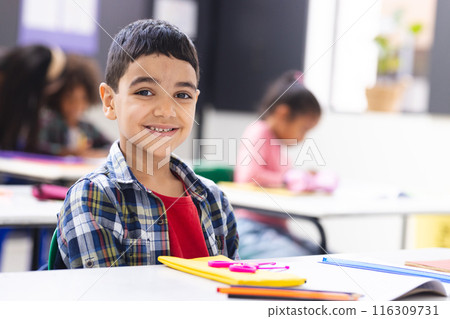 Biracial boy smiling at desk in classroom with school supplies around him 116309731