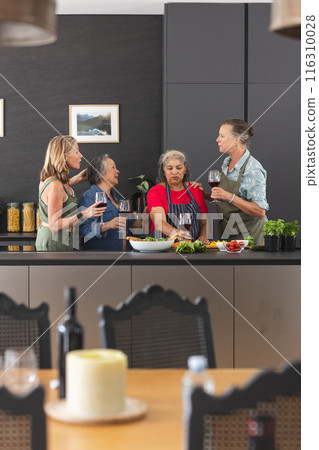 Diverse group of mature women enjoying wine and preparing food together in kitchen 116310028
