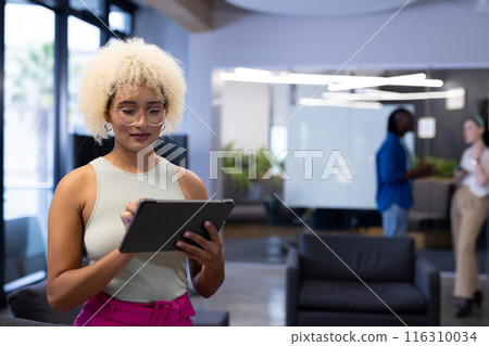 Young biracial woman using tablet at office as colleagues chat in the background, copy space 116310034