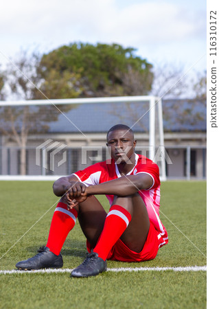 Young African American athlete in red sportswear sitting on soccer field resting 116310172