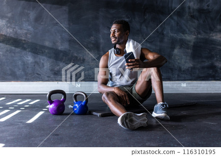 Fit African American man sitting on gym floor with smartphone after exercising, copy space 116310195
