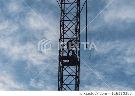 A spectacular silhouette of a man working on a metal tower against a cloudy sky. Loosely hanging cables indicate construction, repair or telecommunication work. A spectacular silhouette of a man working on a metal tower against a cloudy sky. Loosely hanging cables indicate construction, repair or telecommunication work. 116310393