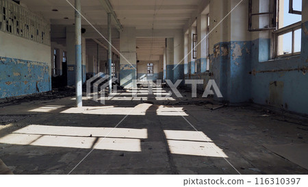An abandoned, dilapidated building with peeling paint and broken windows. Sunlight streams in through the windows, casting shadows on the rubble-strewn floor. The interior of the building is empty. 116310397