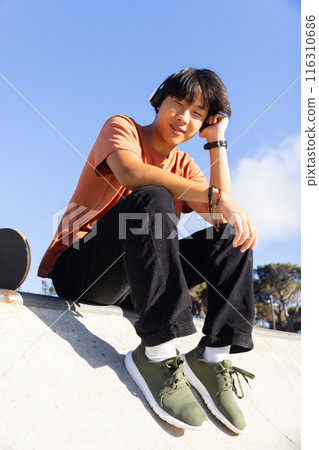 Listening to music with headphones, Asian teenage boy sitting at skatepark 116310686