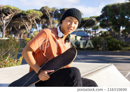 Teenage boy holding skateboard and wearing headphones, relaxing at skate park 116310690