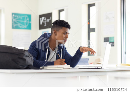 In school, African American male student using laptop and writing in notebook, studying 116310724