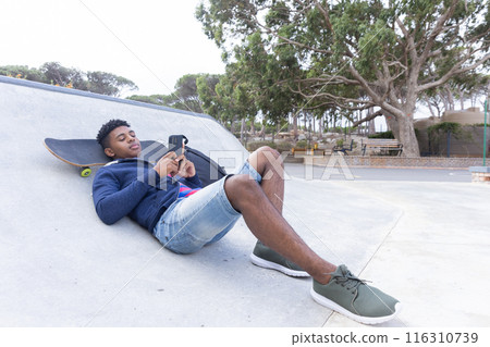 Relaxing at skatepark, African American teenage boy using smartphone, skateboard beside Relaxing at skatepark, African American teenage boy using smartphone, skateboard beside 116310739