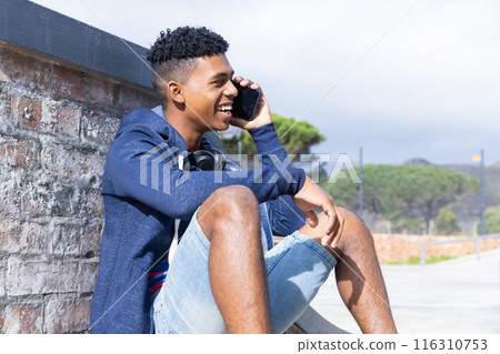 Sitting outdoors, teenage boy talking on smartphone, smiling and enjoying conversation 116310753