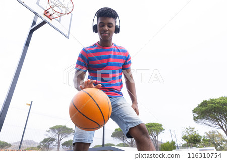 Dribbling basketball, teenage boy wearing headphones enjoying outdoor activity on court 116310754