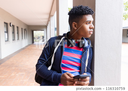 male student holding smartphone and wearing headphones, leaning against pillar in school, copy space male student holding smartphone and wearing headphones, leaning against pillar in school, copy space 116310759