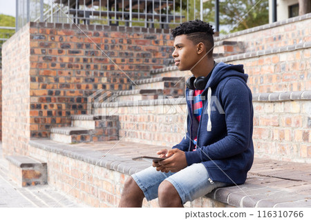 Holding smartphone and wearing headphones, male student sitting on steps relaxing outdoors Holding smartphone and wearing headphones, male student sitting on steps relaxing outdoors 116310766