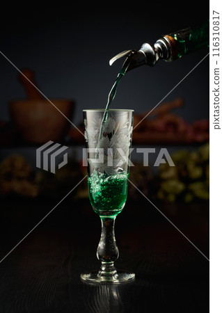 Green herbal liquor is poured from a vintage bottle into a glass. 116310817