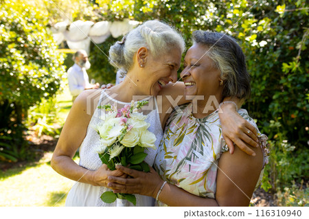 Senior women hugging and smiling, holding bouquet of flowers in garden 116310940