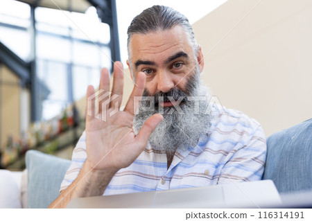 Senior man waving hand while video chatting on laptop at home 116314191