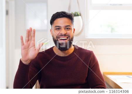 Smiling man waving hand while sitting at home, looking at camera, on video call 116314306