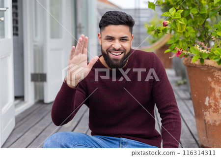 Smiling man waving hand while sitting on wooden deck at home, on video call Smiling man waving hand while sitting on wooden deck at home, on video call 116314311