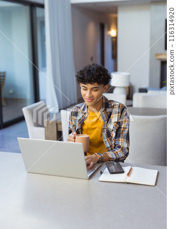 Working from home, teenage boy using laptop and holding coffee mug at table Working from home, teenage boy using laptop and holding coffee mug at table 116314920
