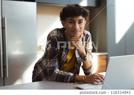 Smiling teenage boy using laptop, leaning on kitchen counter at home 116314941