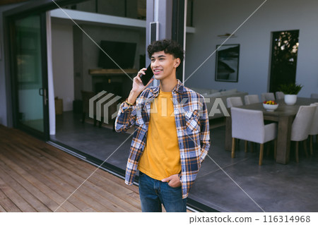 Talking on smartphone, teenage boy standing on patio in casual outfit 116314968