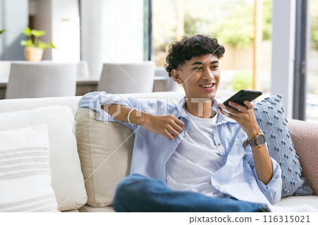 Relaxing on couch, teenage boy using smartphone and smiling at home 116315021