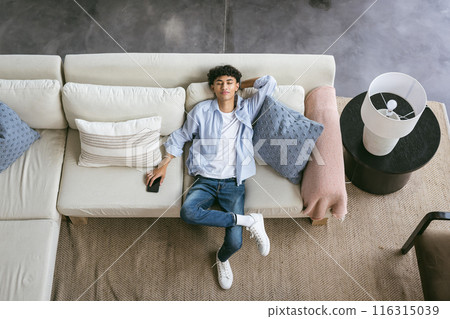 Relaxing on couch, young man holding smartphone and looking up at camera 116315039